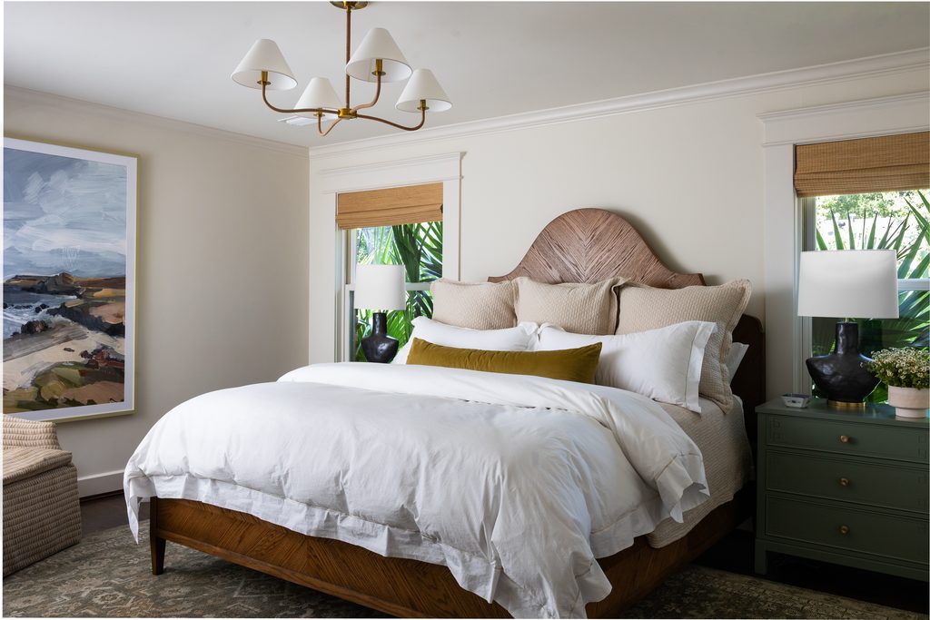 Bedroom with a brass chandelier, black table lamps on the nightstands, woven window shades, and a landscape painting, designed by Elizabeth Ryan Interiors