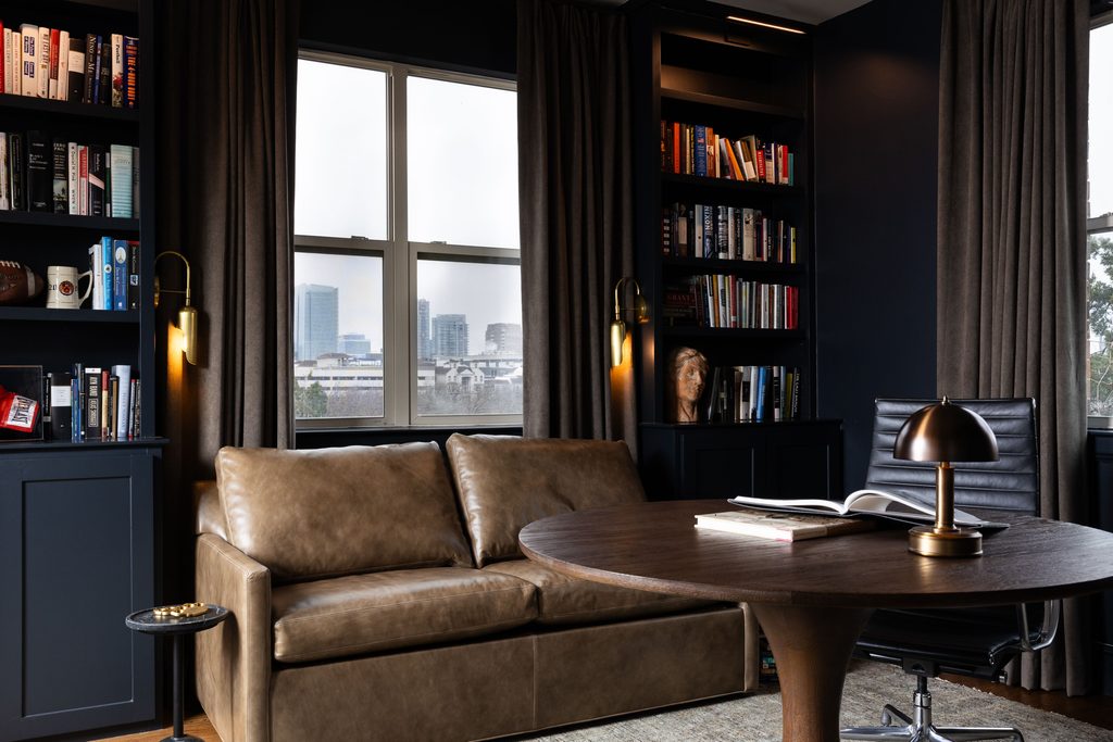 Dark study with built-in bookshelves, brass wall sconces, a leather sofa, and a domed desk lamp, with drapery framing city-view windows