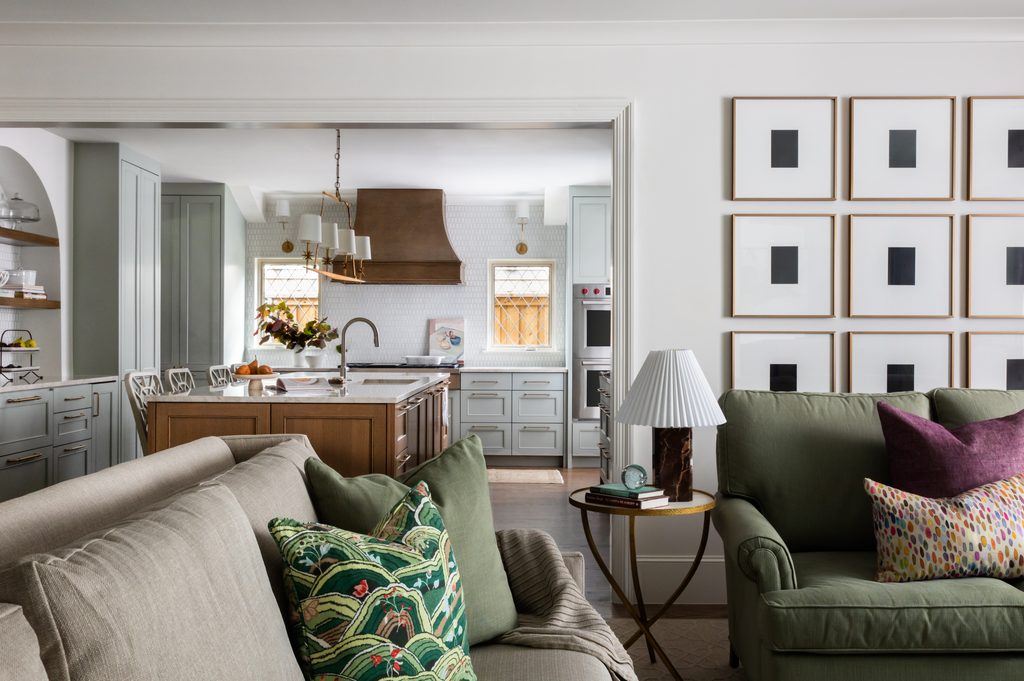 Living room opening into a kitchen with a chandelier over the island, sconces above the windows, and a pleated table lamp beside the sofa